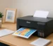 Impressora Epson L3250 em uma mesa de home office imprimindo uma folha colorida com quadrados, ao lado de documentos, caneca e planta decorativa.